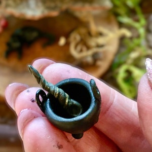 Puede incluir: Un caldero negro en miniatura con un palo de agitación dorado y verde. El caldero es del tamaño de una uña.