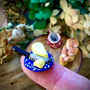 Könnte beinhalten: Ein kleiner blauer Teller mit weißem Rand und weißem Design. Ein kleines Messer und zwei Stücke gelber Knete in Form von Butter befinden sich auf dem Teller.