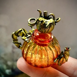 Könnte beinhalten: Eine kleine, dekorative Teekanne mit einem kürbisförmigen Design. Der durchscheinende orangefarbene Körper hat goldene Akzente, mit einem goldenen Deckel und Griff. Diese Miniatur-Teekanne ist wahrscheinlich ein Sammlerstück oder ein Ziergegenstand.