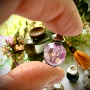 Puede incluir: Una botella de vidrio púrpura en miniatura con una tapa plateada. La botella está siendo sostenida por una mano.