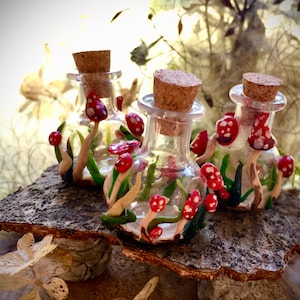 May include: Three small glass bottles with cork stoppers, filled with red and white mushrooms and green foliage. The bottles are sitting on a piece of bark.