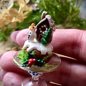 May include: A miniature gingerbread house with white icing and a snowman figurine sits on a glass pedestal with a green moss base and red berries.