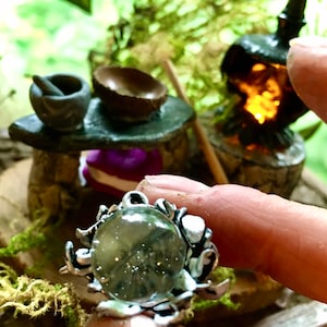May include: A silver ring with a clear glass stone set in a swirling, organic design. The ring is held in a hand, with a miniature scene of a fireplace and a mortar and pestle in the background.