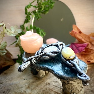 May include: A miniature ceramic table with a light pink jar and a silver and white decorative spoon on top. The table is sitting on a wooden surface.