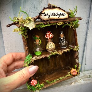 Puede incluir: Un estante de madera en miniatura con un letrero que dice "Titchy Witchy Woo". El estante tiene dos estantes y está decorado con musgo, enredaderas y hongos. Hay tres botellas de vidrio en los estantes, dos de las cuales están llenas de un líquido claro y una de un líquido oscuro.