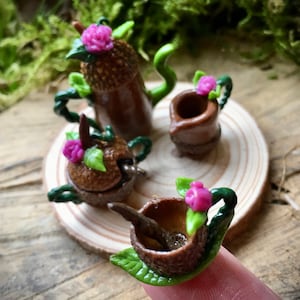 May include: A miniature tea set made from acorns, with a teapot, creamer, sugar bowl, and two teacups. Each piece is decorated with a pink flower and green leaves.