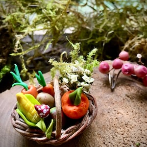 May include: A miniature woven basket filled with small clay vegetables including a yellow corn cob, orange carrots with green tops, a round orange pumpkin, and a tiny red mushroom with white spots, accented with bits of dried green moss and foliage.