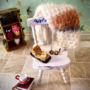 May include: A white painted wooden chair with a fluffy brown and white blanket draped over the back. A small open book with a handwritten note sits on the chair's seat, along with a pair of gold-rimmed glasses. A small white teacup with red polka dots sits on a stack of books.