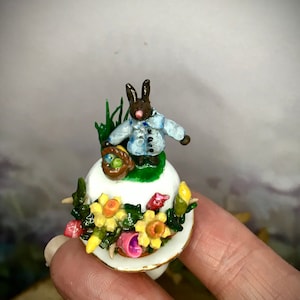 Puede incluir: Una figura en miniatura con temática de Pascua. Un conejito marrón con una chaqueta azul sostiene una cesta de huevos. La figura está sobre una base blanca adornada con flores y vegetación, creando una escena primaveral. La base mide unos 2,5 cm de diámetro.