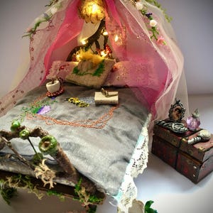May include: A miniature fairy bed with a grey bedspread, embroidered details, and a pink canopy. The bed is decorated with small pillows, a teacup, and a decorative chest. The bed frame is made of wood and moss.