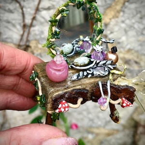 Puede incluir: Un conjunto de tocador en miniatura hecho de madera y decorado con hiedra, champiñones y cristales. El tocador tiene un pequeño espejo, un tarro rosa y una bandeja plateada con cristales y una botella.