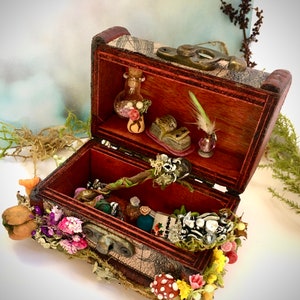 May include: A small, open wooden treasure chest filled with miniature items like bottles and a key. The chest is adorned with moss, flowers, and a mushroom. The interior is lined with patterned fabric, and the wood is stained a dark brown colour.