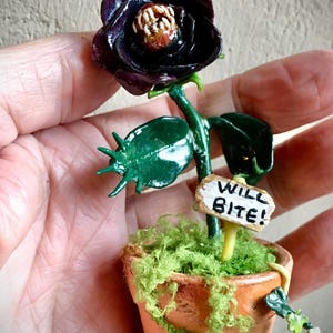 May include: A miniature, handcrafted, dark purple rose with a detailed center, set in a small terracotta pot. The pot is filled with green moss and has a small sign that reads "WILL BITE!"