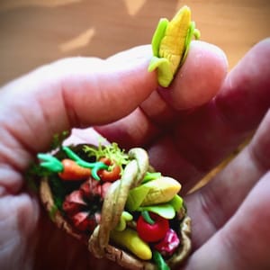 May include: A miniature basket filled with a variety of colorful vegetables, including corn, carrots, and tomatoes. The basket is made of brown and gold clay and has a rustic, handmade look. A small corn cob is held in a hand, showcasing the miniature scale of the items.