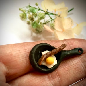 Puede incluir: Una sartén miniatura negra con una salchicha marrón y un huevo frito dentro. La sartén tiene un mango negro.