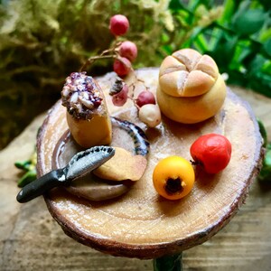 May include: A miniature wooden table with a round top and a single leg. The table is set with a small loaf of bread, a knife, a plate with a piece of bread, and two small fruits.