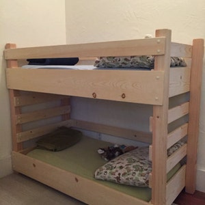 May include: A wooden bunk bed with a natural finish. The bottom bunk has a green mattress and a patterned pillow. The top bunk has a blue mattress and a patterned pillow.