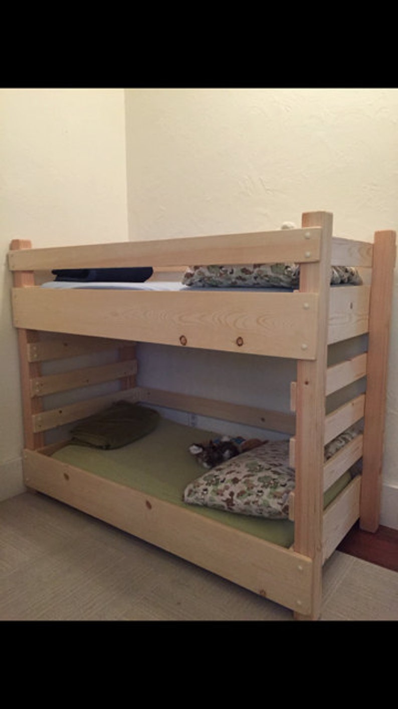 May include: A wooden bunk bed with a natural finish. The bottom bunk has a green mattress and a patterned pillow. The top bunk has a blue mattress and a patterned pillow.