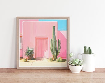 May include: A framed painting of a pink building with a pink door and a cactus in front of it. The building is set against a blue sky.