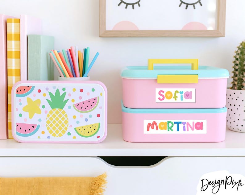 May include: Two rectangular pink lunch boxes with light blue lids. One box features a pineapple, watermelon slices, and colorful dots. The other stacked box has a yellow handle and two white labels with the names Sofia and Martina in colorful letters.