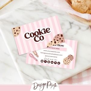 Può includere: Due biglietti da visita a righe rosa e bianche con la scritta "Cookie Co" in marrone. I biglietti presentano illustrazioni di biscotti e informazioni di contatto. Sono su una superficie di marmo, con prodotti da forno sullo sfondo.