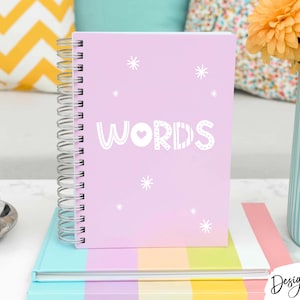 May include: A light pink spiral-bound notebook featuring the word WORDS printed in white capital letters with a small heart inside the letter O, surrounded by white starburst graphics on the front cover, resting on a multi-colored striped book.
