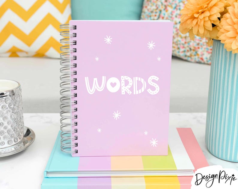 May include: A light pink spiral-bound notebook featuring the word WORDS printed in white capital letters with a small heart inside the letter O, surrounded by white starburst graphics on the front cover, resting on a multi-colored striped book.