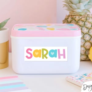 May include: A rectangular white plastic storage container with a light pink lid featuring a colorful leaf pattern. The front of the container displays a white rectangular label with the name Sarah printed in large, multicolored, patterned letters.
