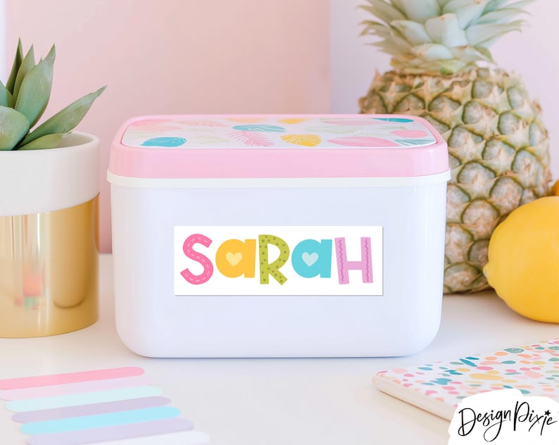 May include: A rectangular white plastic storage container with a light pink lid featuring a colorful leaf pattern. The front of the container displays a white rectangular label with the name Sarah printed in large, multicolored, patterned letters.