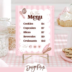 May include: A pink and white menu sign with a gold stand displays a bakery's offerings: cookies, cupcakes, slices, brownies, and croissants. The sign features a cupcake and spoon illustration, with the text "On The Menu" in brown.