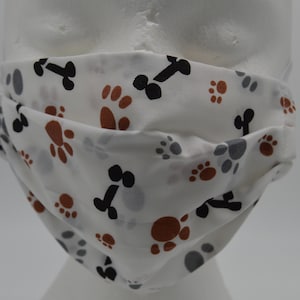Op de afbeelding: Witte gezichtsmasker met een herhalend patroon van bruine poot afdrukken, zwarte botvormen en grijze poot afdrukken.