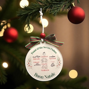 Puede incluir: Una bola de Navidad redonda y transparente con el texto "Feast of the Seven Fishes" y "Buon Natale". El adorno presenta ilustraciones de mariscos y está adornado con un lazo a rayas rojo y verde y una cuenta roja. Cuelga de un árbol de Navidad.