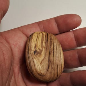 May include: A small, oval-shaped wooden object with a natural wood grain pattern. The wood has a light brown color with darker brown streaks, and a smooth, polished finish. The object is held in a hand.