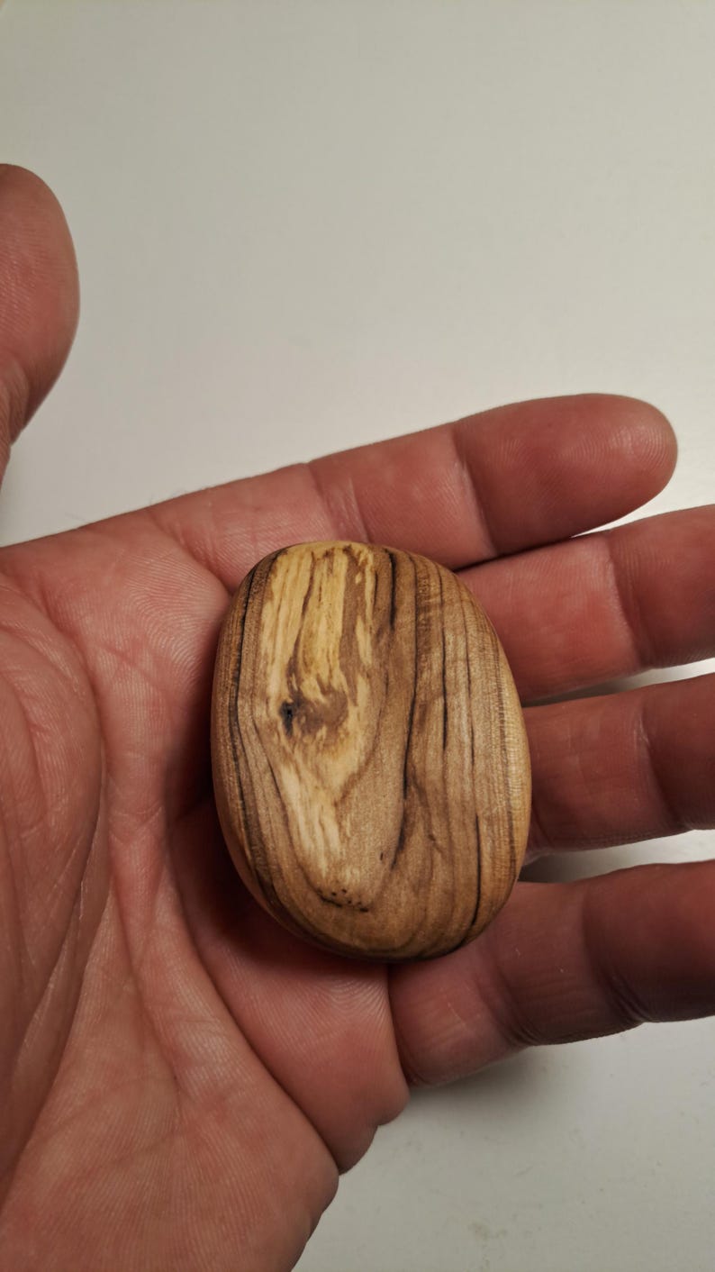 May include: A small, oval-shaped wooden object with a natural wood grain pattern. The wood has a light brown color with darker brown streaks, and a smooth, polished finish. The object is held in a hand.