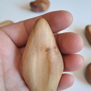 May include: A light-colored, wooden, diamond-shaped object is held in a hand. The wood grain is visible, and the object has a smooth, polished surface. Other similar wooden objects are in the background.