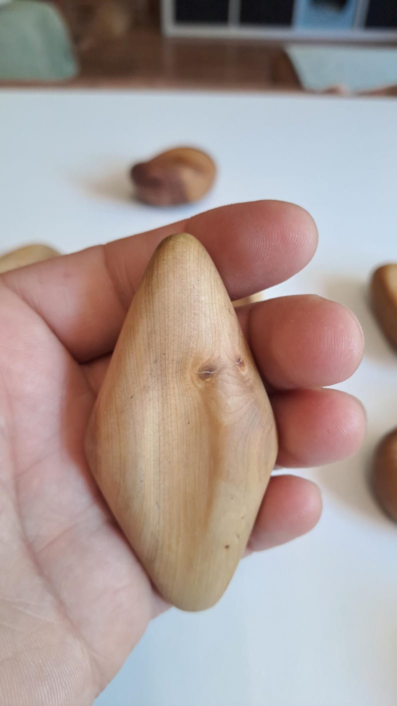 May include: A light-colored, wooden, diamond-shaped object is held in a hand. The wood grain is visible, and the object has a smooth, polished surface. Other similar wooden objects are in the background.
