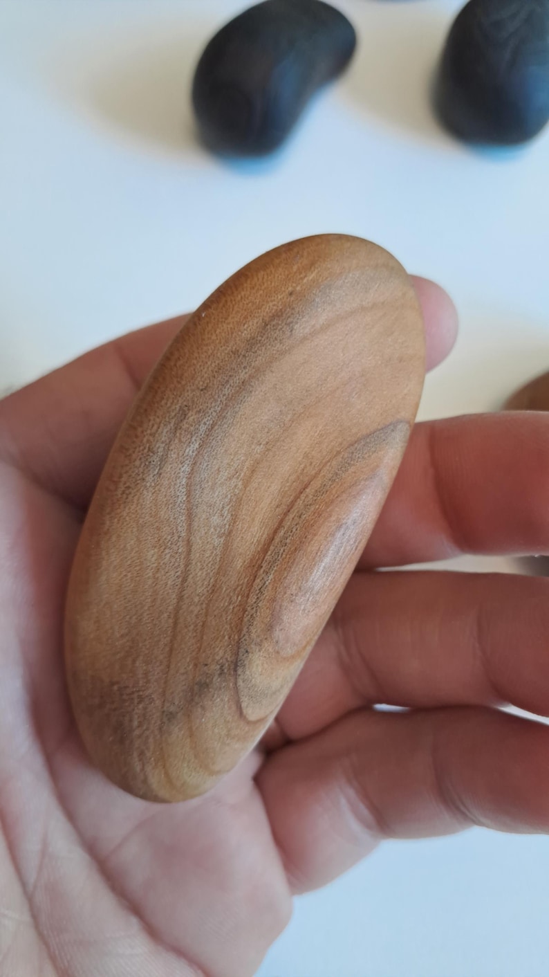 May include: A light brown, oval-shaped wooden object with visible wood grain. The object is held in a hand, with other dark-colored, pebble-like objects visible in the background. The wood has a natural, warm tone.