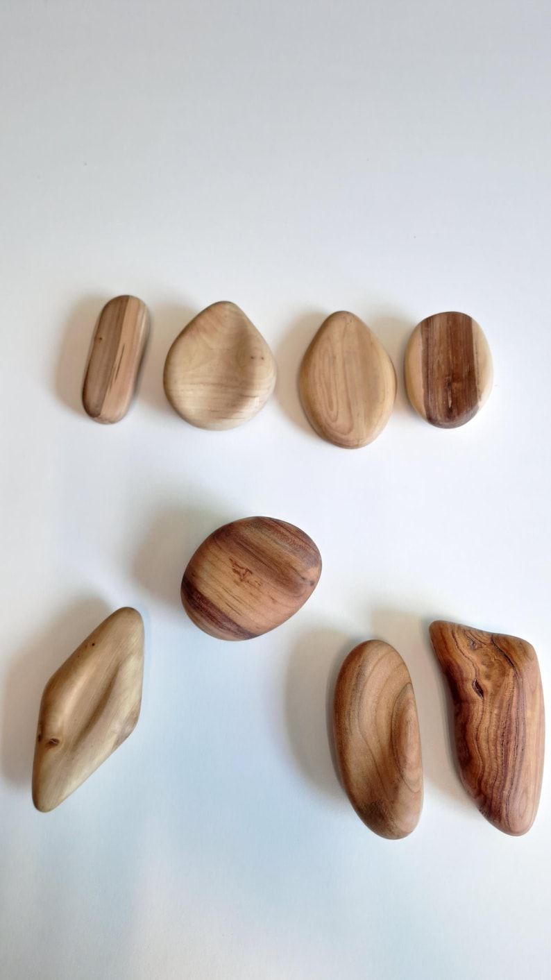 May include: A collection of eight smooth, wooden stones in various shapes and sizes. The stones are light brown with natural wood grain patterns. Some are oval, others are more angular, and all are set against a white background.