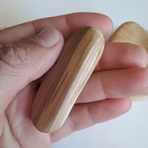 May include: A light-colored, wooden massage tool with a rounded shape, held in a hand. The wood grain is visible, showing a natural pattern. Another wooden piece is partially visible in the background.