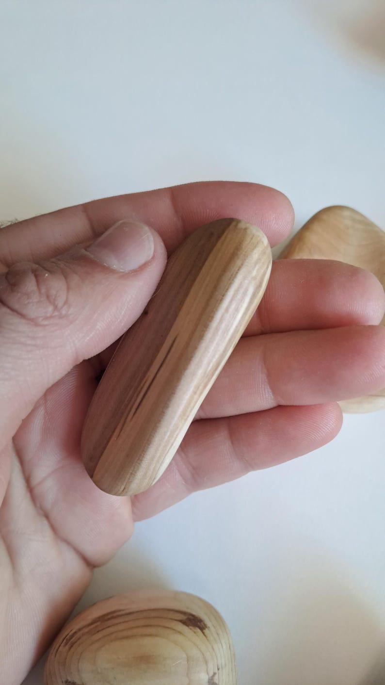 May include: A light-colored, wooden massage tool with a rounded shape, held in a hand. The wood grain is visible, showing a natural pattern. Another wooden piece is partially visible in the background.