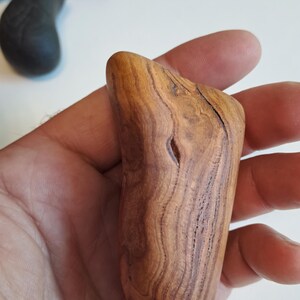 May include: A hand-held, smooth, wooden massage tool with a natural wood grain pattern. The tool is a light brown color with darker brown streaks and a small hole. Other wooden objects are in the background.