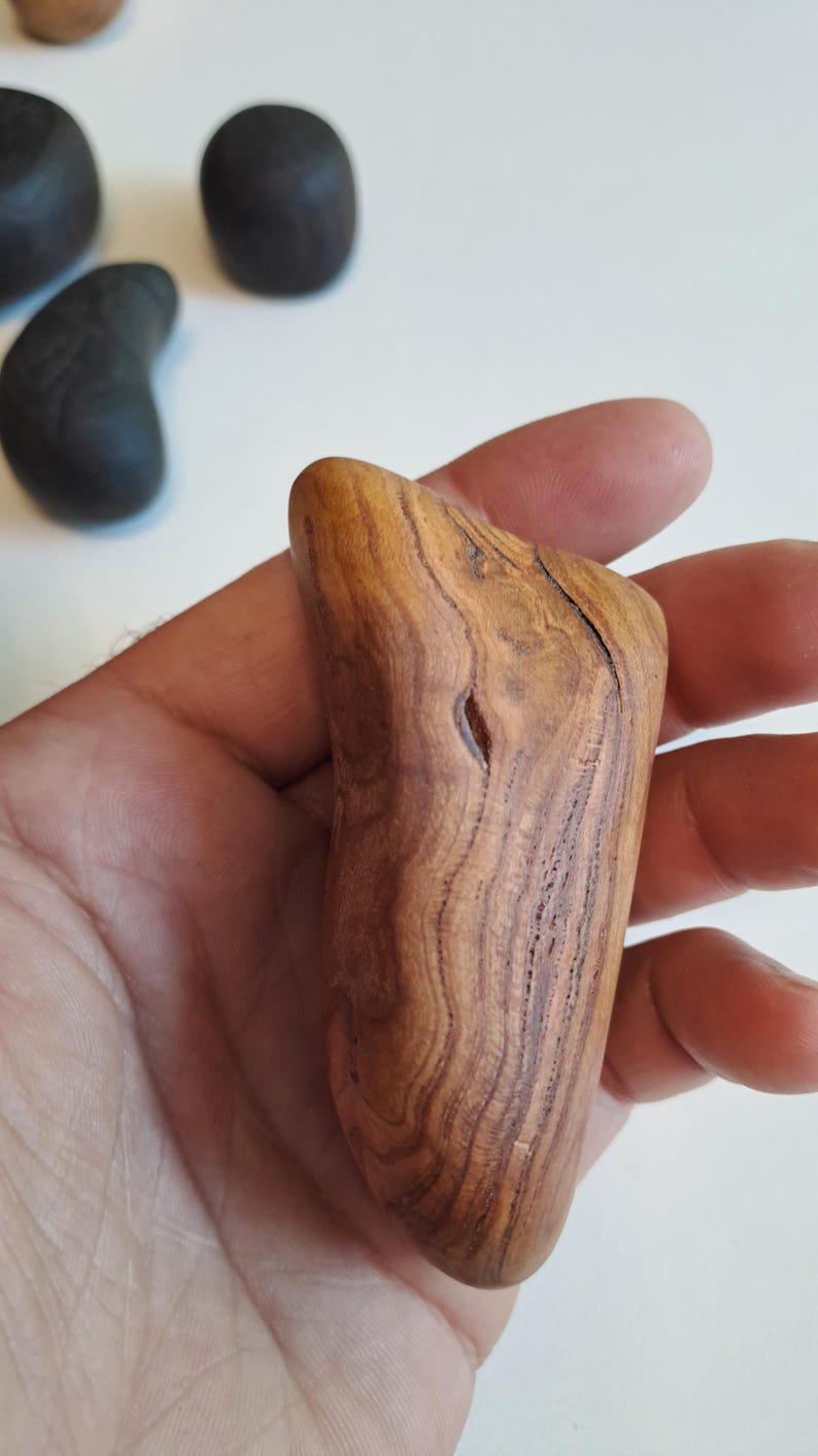 May include: A hand-held, smooth, wooden massage tool with a natural wood grain pattern. The tool is a light brown color with darker brown streaks and a small hole. Other wooden objects are in the background.