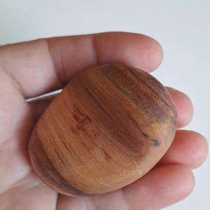 May include: A smooth, oval-shaped wooden stone with a natural wood grain pattern. The stone is a warm brown color with darker brown streaks and a polished finish. The stone is held in a hand.