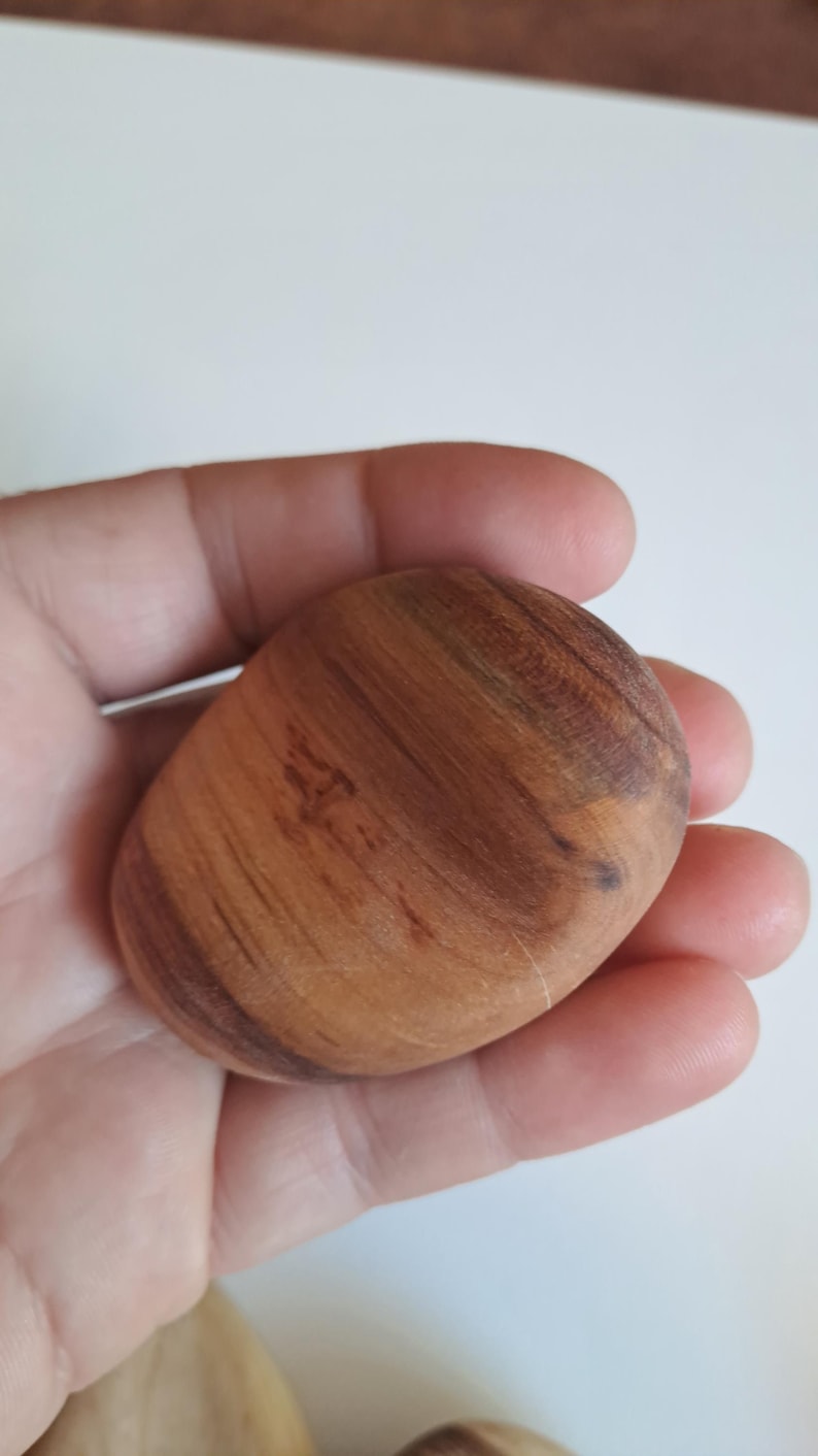 May include: A smooth, oval-shaped wooden stone with a natural wood grain pattern. The stone is a warm brown color with darker brown streaks and a polished finish. The stone is held in a hand.