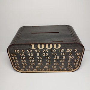 May include: A wooden money-saving box with a dark gray finish. The box has a slot on top and a front panel with the number "1000" and a grid of numbers. The box is designed for saving money.