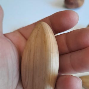 May include: A light-colored, teardrop-shaped wooden object is held in a hand. The wood grain is visible, and the object has a smooth, polished appearance. Other similar wooden objects are visible in the background.