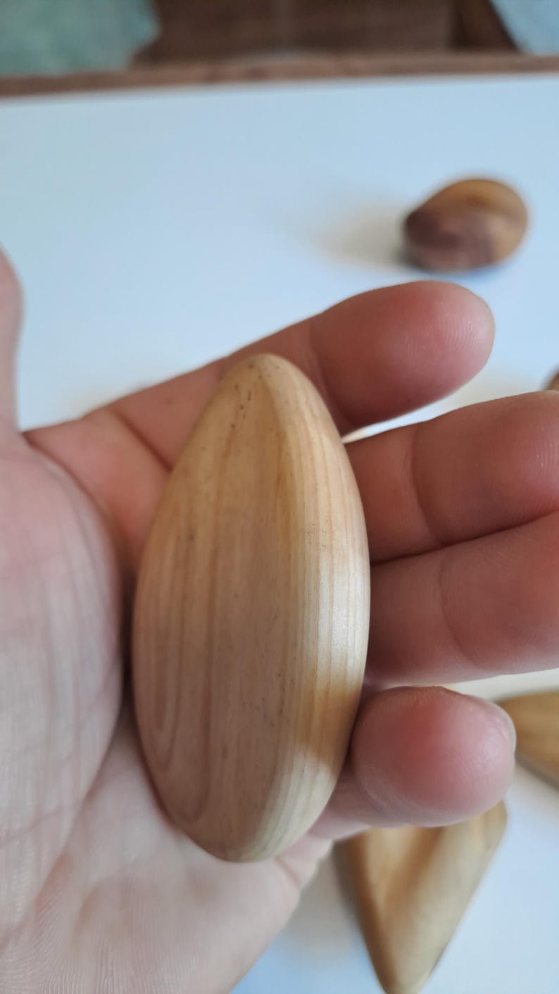 May include: A light-colored, teardrop-shaped wooden object is held in a hand. The wood grain is visible, and the object has a smooth, polished appearance. Other similar wooden objects are visible in the background.