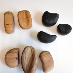 May include: An assortment of wooden massage tools in various shapes and sizes. Some are light-colored with visible wood grain, while others are dark charcoal-colored. The tools are arranged on a white surface, showcasing their natural textures.