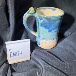 May include: A handmade ceramic mug with a blue and tan glaze, featuring a unique handle design. The mug has the word "Earth" written on a small, rectangular ceramic tile. The mug is set against a dark background.