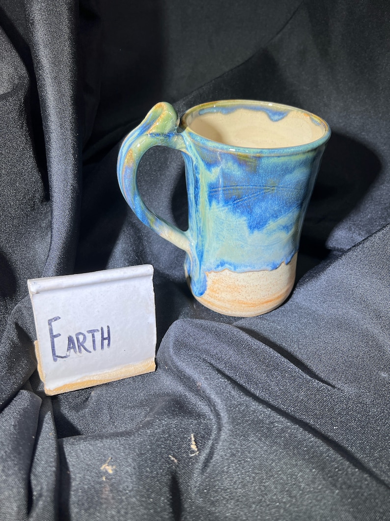 May include: A handmade ceramic mug with a blue and tan glaze, featuring a unique handle design. The mug has the word "Earth" written on a small, rectangular ceramic tile. The mug is set against a dark background.